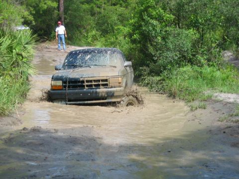 1992 Ford Bronco - A truck is stuck in a muddy puddle on a dirt road, with a person standing in the background.
