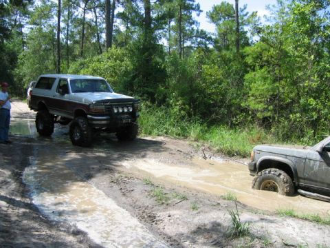1992 Ford Bronco - A lifted 1992 Ford Bronco navigates a muddy off-road trail, with another vehicle struggling in the mire.