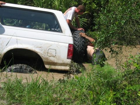 1992 Ford Bronco - Two people attempt to free a white Ford Bronco that is stuck deep in the mud, with one person submerged in the mire.