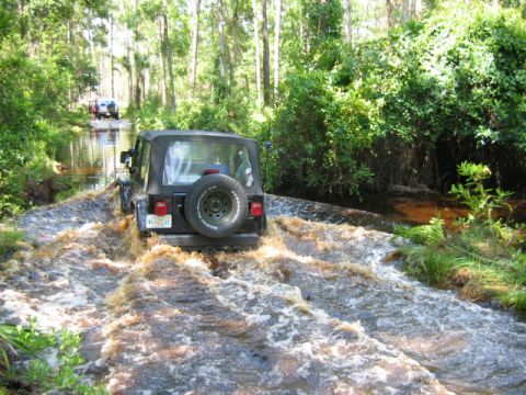 1992 Ford Bronco - Off-roading Jeep Wrangler navigates a water crossing in a wooded area.
