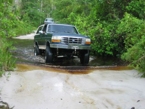 1992 Ford Bronco - Two SUVs navigate a water crossing on a sandy, wooded trail.