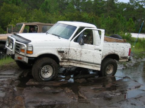 1992 Ford Bronco - A white Ford Bronco is stuck in a muddy off-road course, with another vehicle visible in the background.
