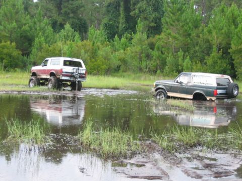 1992 Ford Bronco - Two Ford Broncos navigate a muddy off-road course, with one vehicle partially submerged in water.