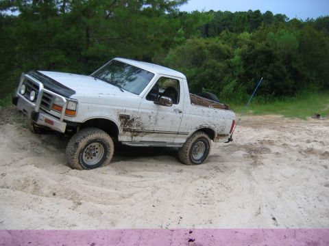 1992 Ford Bronco - White 1992 Ford Bronco navigates a sandy terrain with trees in the background.