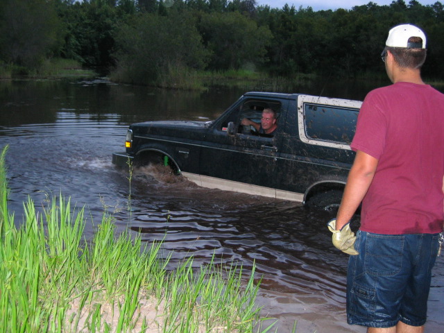 Dark green doorless Ford Bronco driving through a muddy pond with a spotter watching