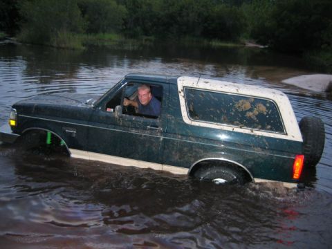 1992 Ford Bronco - A man drives a muddy Ford Bronco through a body of water surrounded by trees.