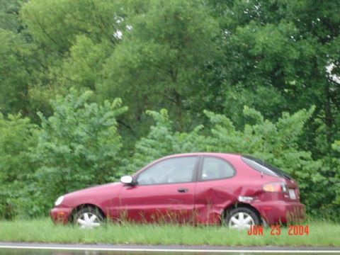 1986 Ford Bronco - Red sedan with visible damage to its rear passenger side quarter panel, parked beside a tree line.