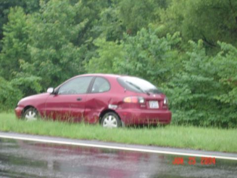 1986 Ford Bronco - A red hatchback car with visible damage to its rear side is parked on the shoulder of a road.