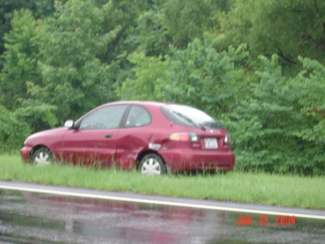 Red hatchback car with damage to rear quarter panel, parked on grassy shoulder next to a road