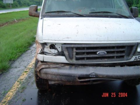 1986 Ford Bronco - A white van with significant front-end damage sits on the side of a wet road.