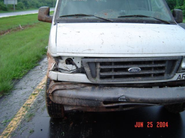 Damaged white van front end, missing headlight, dented bumper, parked on wet roadside