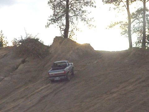 1989 Ford Bronco - Distant rear view of a topless lifted 4x4 climbing a steep dirt hill in a backlit pine-treed dirt pit. The vehicle is silhouetted against a…