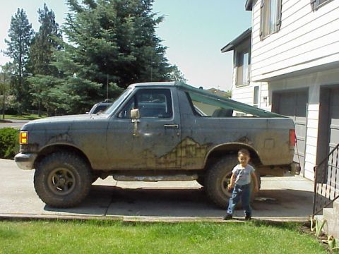 1989 Ford Bronco - A mud-splattered two-door Ford Bronco in primer gray with mountain-scene graphics on the rear quarter, parked in a driveway with the hardto…