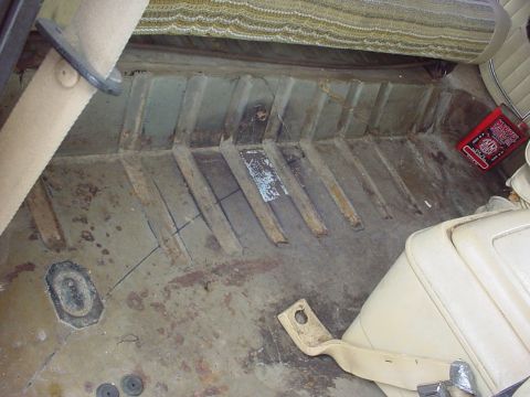 1979 Ford Bronco - The interior floor of a vehicle shows signs of rust and wear, with a red book and beige seats visible.