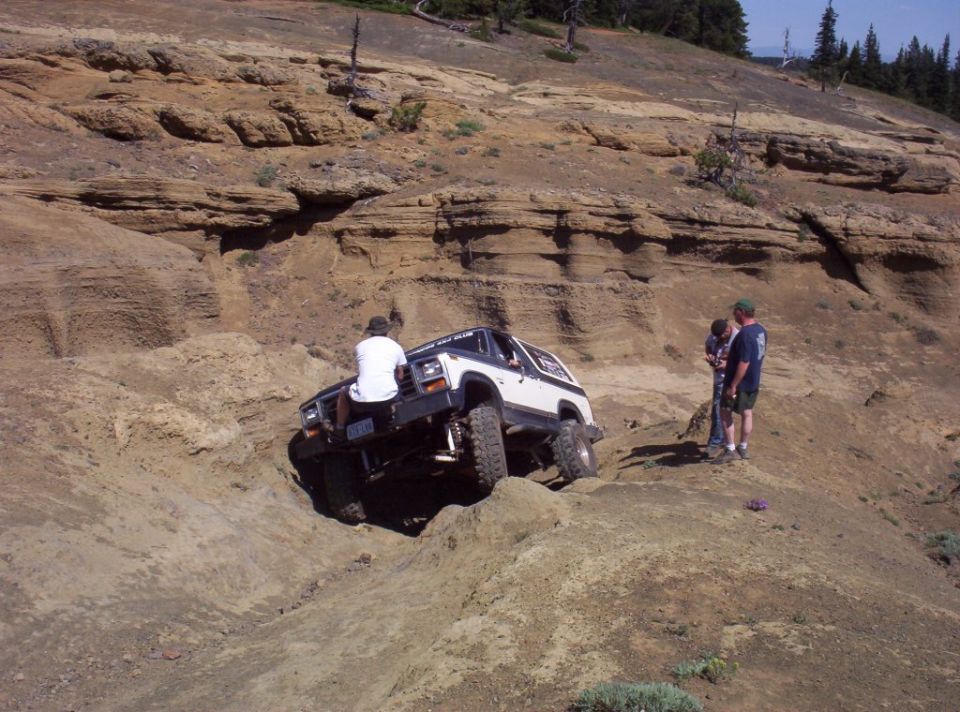 White Bronco tilted in a rock hole on a trail with three spotters nearby