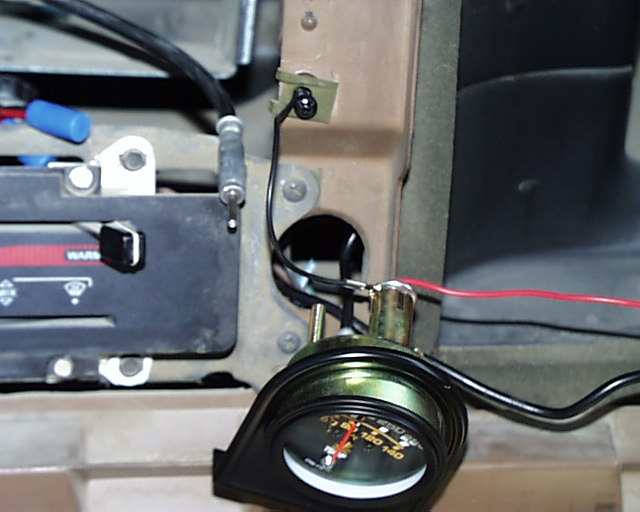Aftermarket round gauge with brass body wired in behind a disassembled truck dash