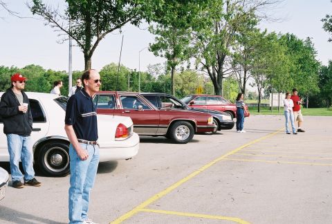 1998 Ford Crown Victoria - Several cars are parked in a lot with people standing around them.