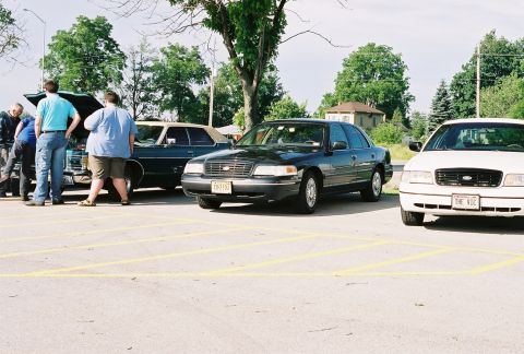 1998 Ford Crown Victoria - Two Ford Crown Victorias are parked in a lot, with people gathered around one of them.