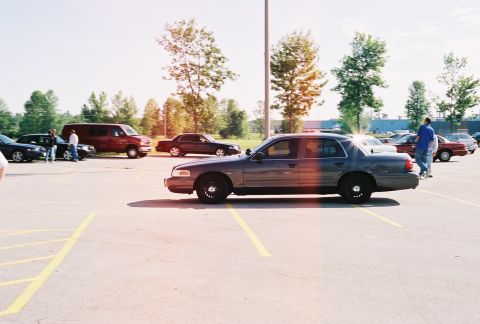 1998 Ford Crown Victoria - A dark sedan is parked in a lot with other vehicles and trees in the background.