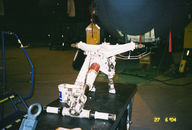 Aircraft landing gear assembly on a table, with a blue cart and a tire visible