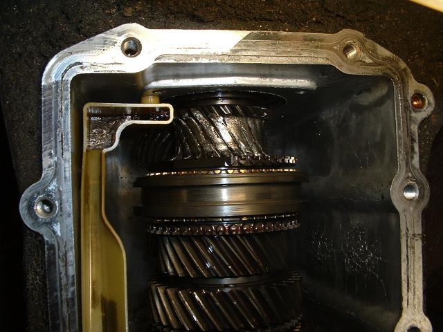 Open manual transmission case showing helical gears, synchro rings, and plastic oil trough inside aluminum housing