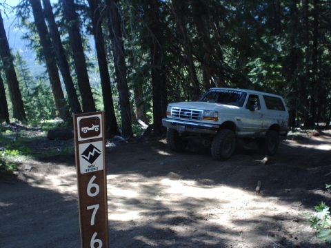 1978 Ford Bronco - A white 1996 Ford Bronco is parked on a dirt trail in a wooded area, next to a trail marker indicating difficulty.