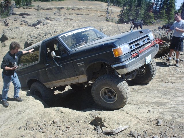 Black Ford Bronco with lifted suspension navigating a rocky trail. A person stands by the driver's door, another person and a dog are in the background