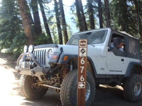 1978 Ford Bronco - A white Jeep Wrangler is parked on a dirt trail surrounded by trees.