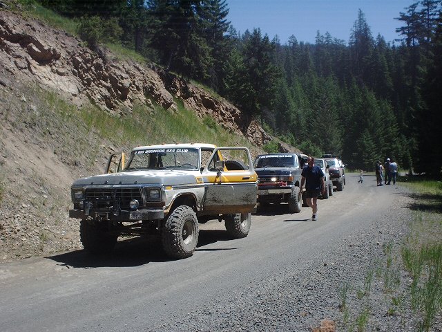 1978 Ford Bronco with yellow and grey paint, lifted suspension, and off-road tires, driving on a gravel road