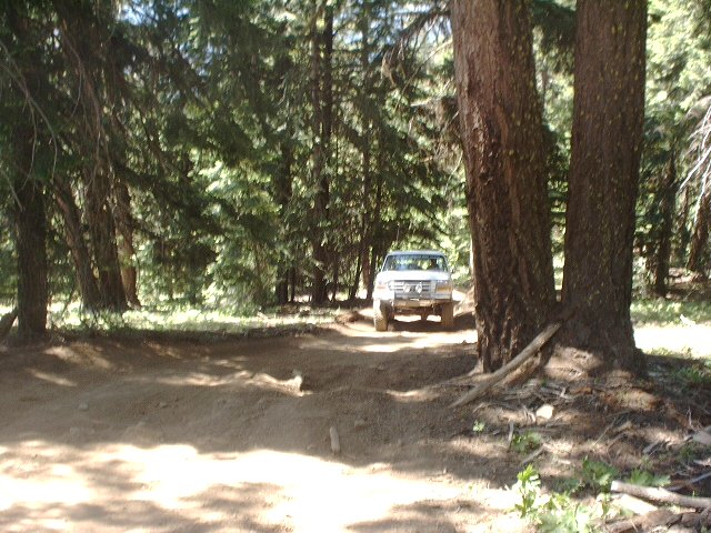 White SUV on a dirt road in a forest. The vehicle has a boxy body and a prominent grille