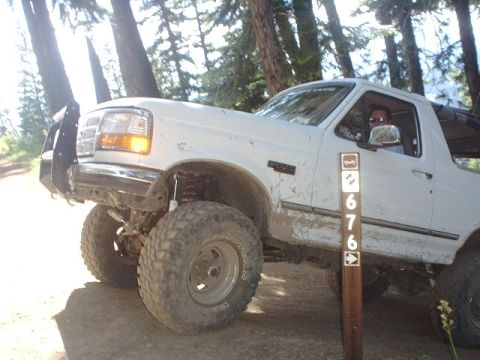 1978 Ford Bronco - White Ford Bronco with large tires and brush guard on a dirt road, driver visible.