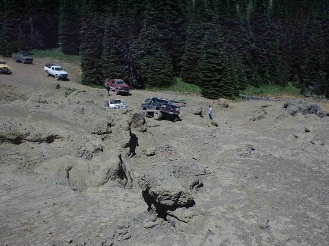 Dark SUV with a lifted suspension navigates a steep, rocky incline, with other trucks and SUVs visible on the trail