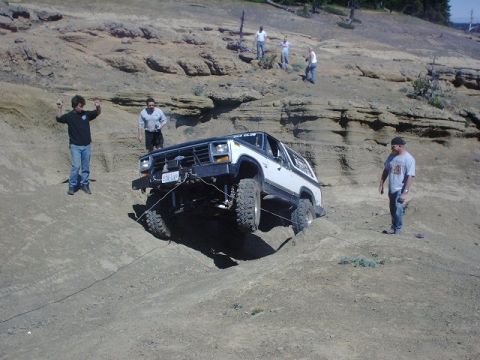1978 Ford Bronco - An off-road SUV navigates a steep, rocky terrain with assistance from onlookers and a winch line.