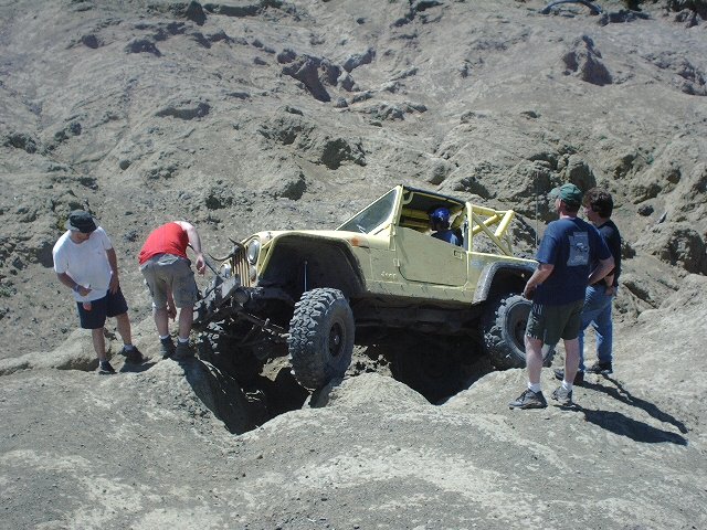 Yellow Jeep Wrangler modified for off-roading, stuck on a rocky incline with people around it