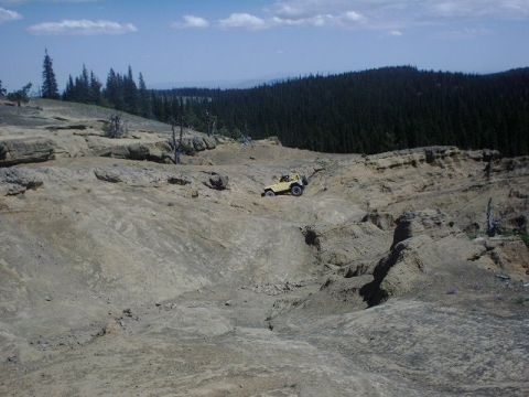 1978 Ford Bronco - A yellow Jeep navigates a , rocky terrain under a blue sky with scattered clouds.