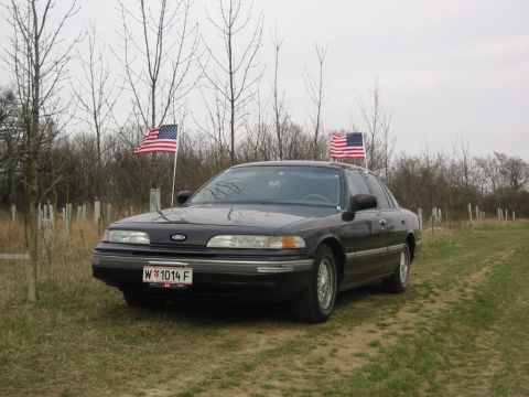 1992 Ford Crown Victoria - A 1992 Ford Crown Victoria sedan is parked outdoors with American flags attached to its sides.