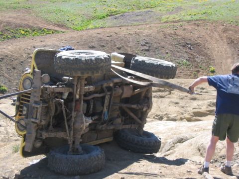 1995 Ford Bronco - A yellow off-road vehicle has overturned on a rocky, dirt terrain, with a person attempting to assist.