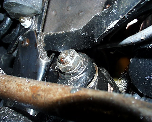 Dark underbody close-up of hex bolt in cast housing with black brackets and rusty tube