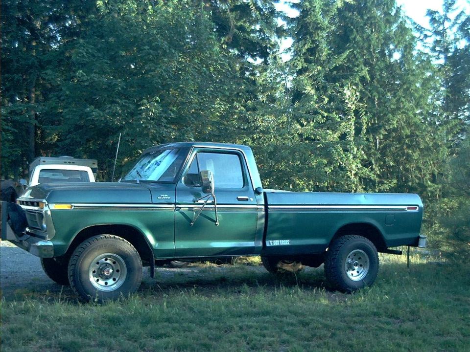Green Ford F-series pickup with white camper shell parked on grass in front of evergreens