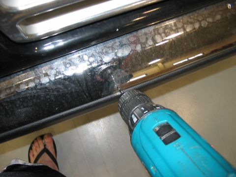 1988 Ford Bronco - A teal power drill is used to drill into a dirty car door sill, with a foot in a flip-flop visible in the lower left corner.