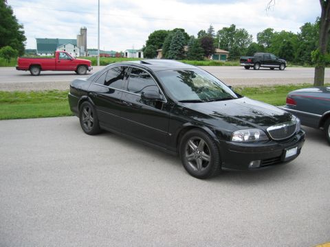 2000 Ford Crown Victoria - Three-quarter front passenger-side view of a black Lincoln LS sedan with tinted windows, chrome waterfall grille and dark five-spoke alloy…