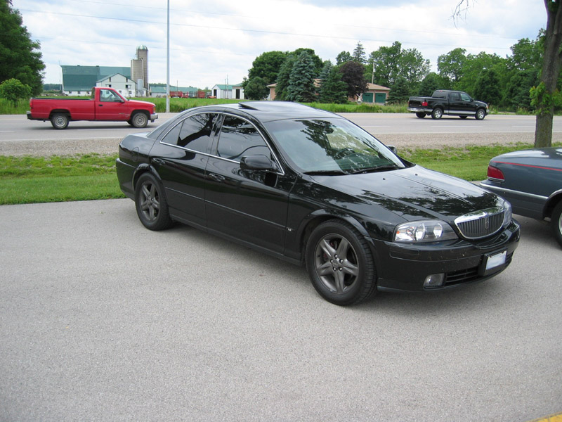 Black Lincoln LS sedan, 3/4 front view, dark alloy wheels, parked on concrete near a rural road