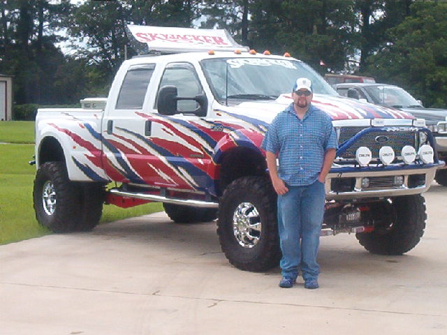 guy standing next to a lifted white pickup truck with red and blue flame graphics and a 'SkyJacker' sign on top