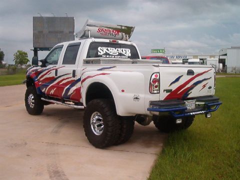 1980 Ford Bronco - Lifted white Ford Super Duty crew-cab dually pickup photographed from the rear three-quarter on a concrete lot, wearing red, white and blue…