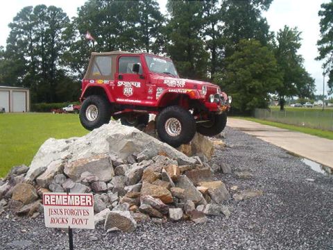 1980 Ford Bronco - A red Jeep Wrangler TJ with a tan soft top and Skyjacker sponsor decals flexes up a rock pile on a gravel lot, riding on beadlock-style whe…