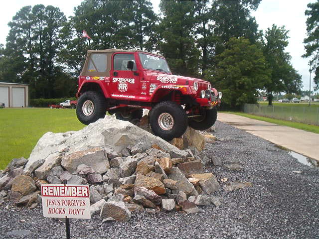 Red Jeep Wrangler with tan soft top climbing a rock pile on a gravel lot