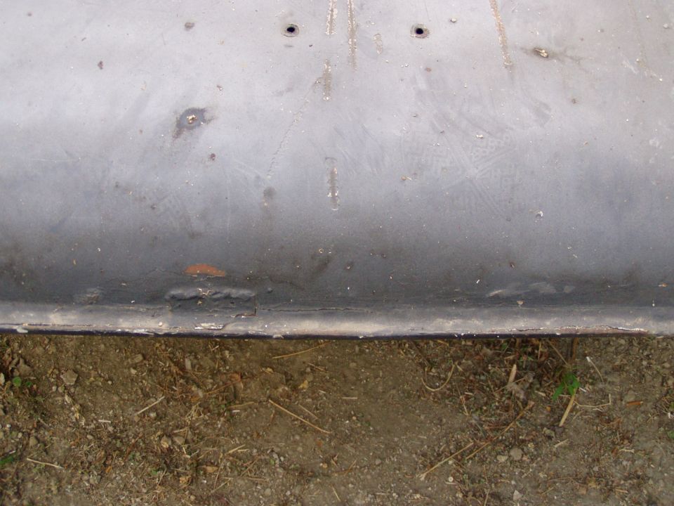Gray metal panel with two holes and visible welds, resting on dirt
