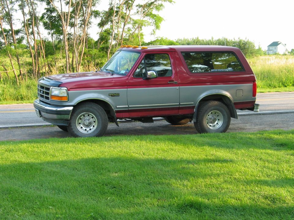 Red and silver two-tone Ford Bronco parked roadside on grass with trees behind