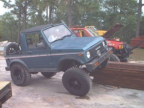 1986 Ford Bronco - A teal Suzuki Samurai is positioned on a stack of lumber, with other vehicles visible in the background.