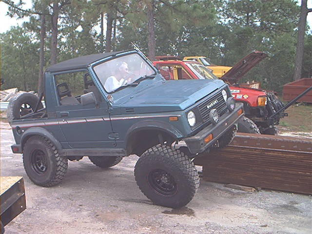 Teal Suzuki Samurai on oversized tires climbing onto stacked steel beams